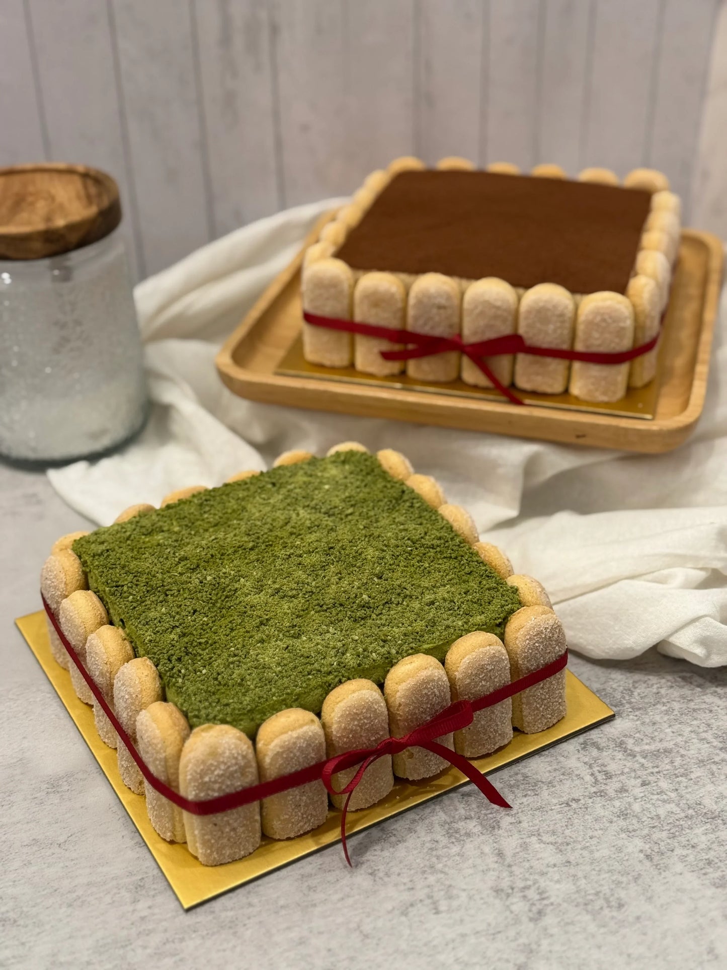 Two square Tiramisu cakes in matcha and classic flavours on a gray surface.