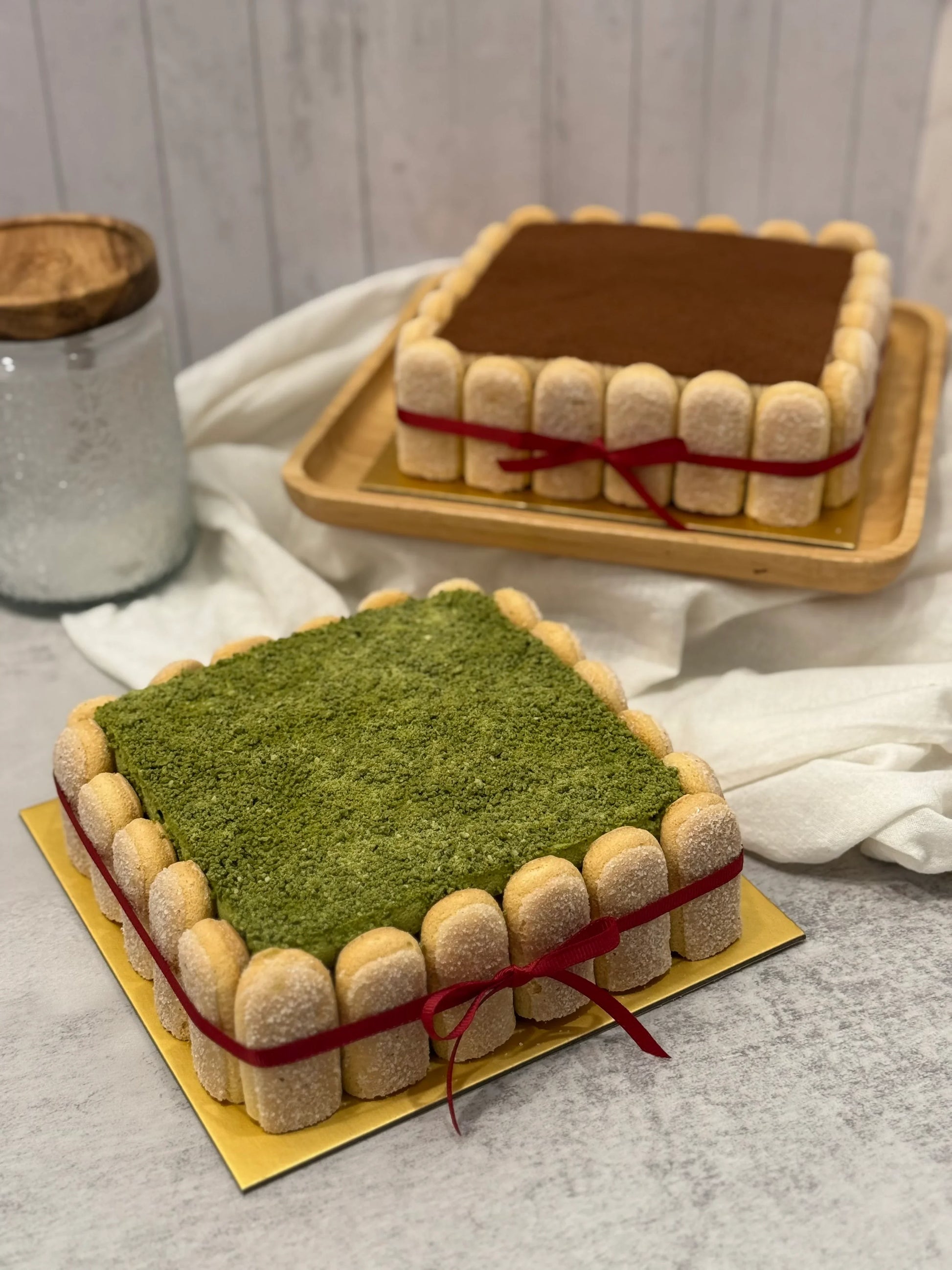 Two square Tiramisu cakes in matcha and classic flavours on a gray surface.