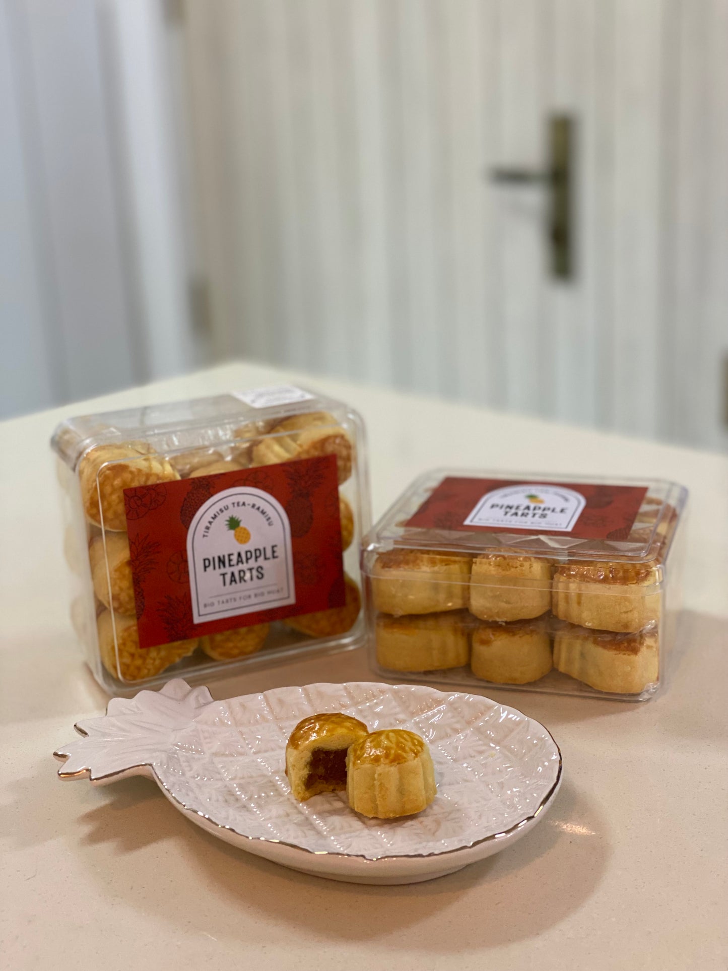 Two containers of pineapple tarts on a table with a decorative plate featuring a pineapple tart cut in half.