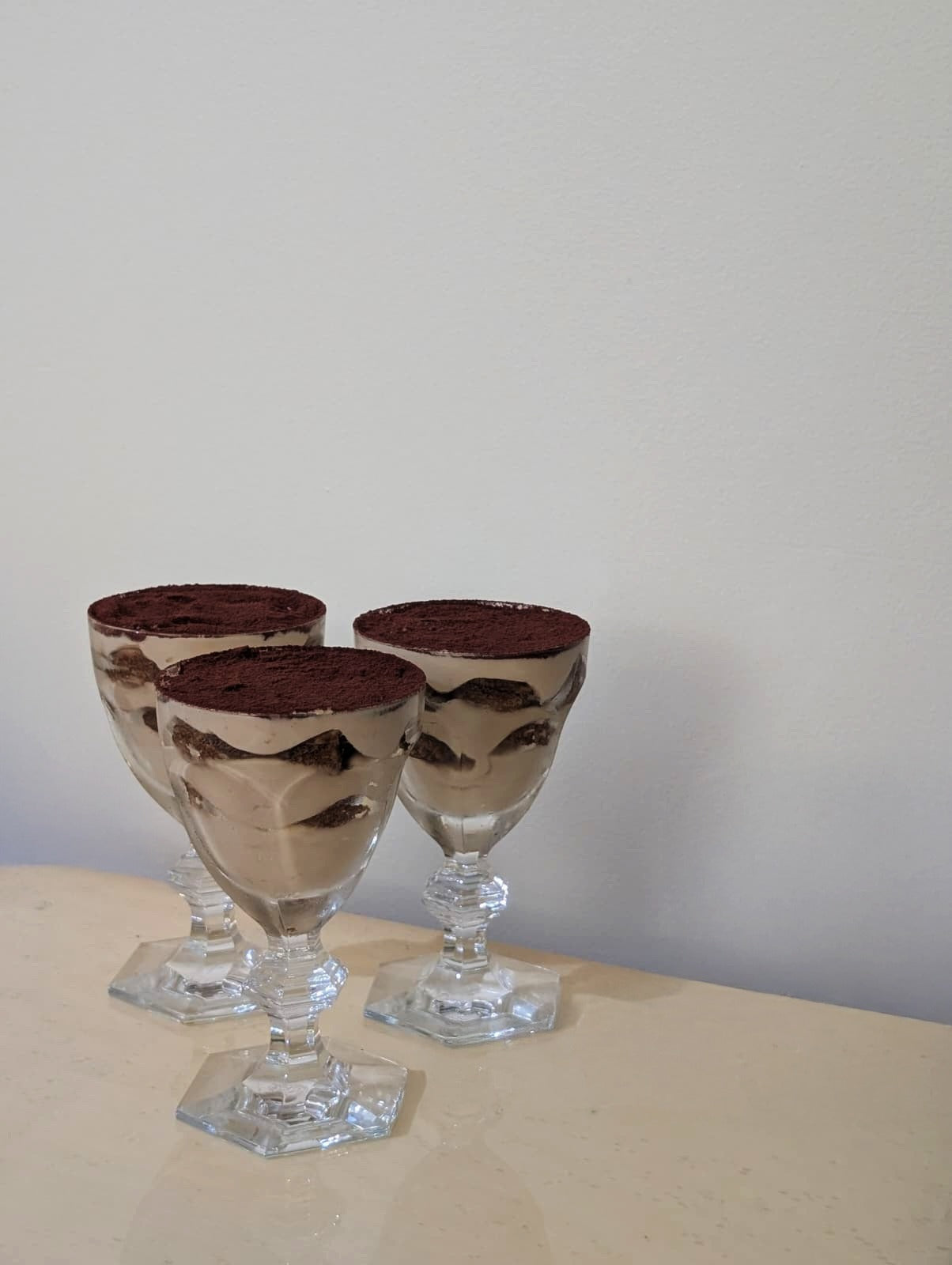 Three glass dishes with a Tiramisu layered dessert on a beige surface and light gray background